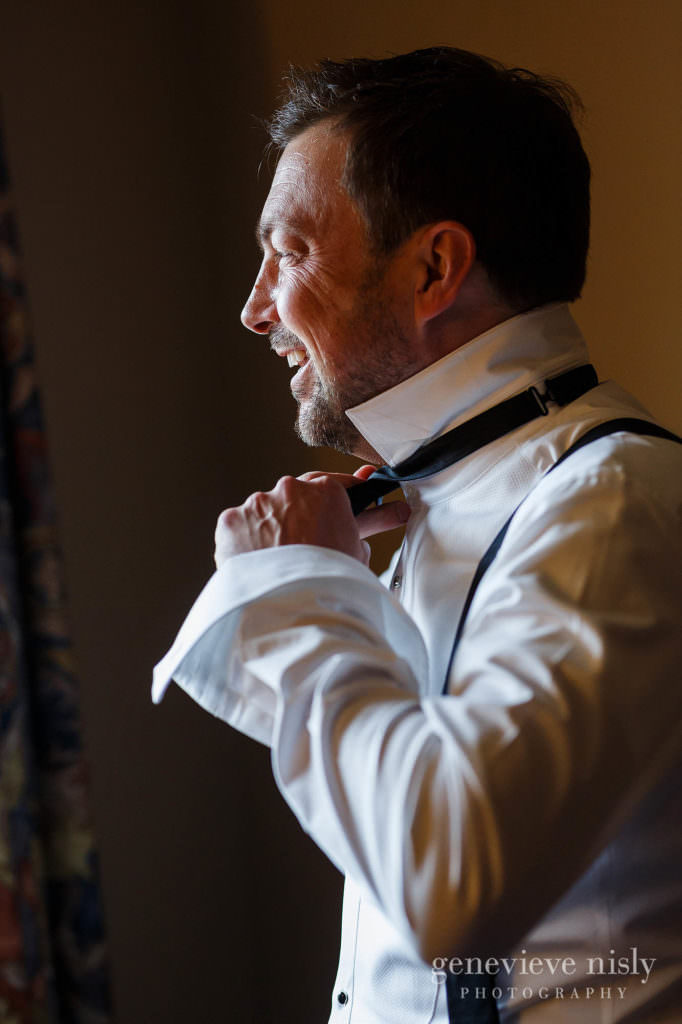 A groom adjusts his bowtie while laughing.