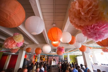 A bride and groom facing each other at the altar during the wedding ceremony at 78TH STREET Studio in Cleveland, Ohio decorated with white and orange paper lanterns and paper flowers hanging from the ceiling.