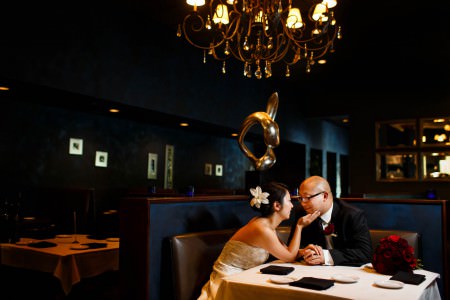 An image of a bride and groom sitting down in a booth at 91 Wood Fired Oven where the bride is wearing a strapless dress and a flower in her updo leaning in towards the groom with her hand touching his chin while the groom is wearing his tuxedo and glasses with a gold chandelier and gold decorations set inside a dark blue room.