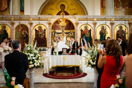 A image of a bride and groom holding candles circling the altar with the priest during a wedding ceremony inside the beautifully ornate Annunciation Greek Orthodox Church with colorful paintings and ornate stone pillars with gold trim throughout the sanctuary.
