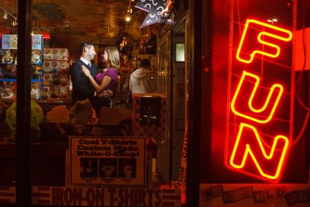 A picture of an engaged couple standing just to the left of the center of the photo facing each other in an embrace while standing in the middle of the Big Fun Toy Store full of trinkets and brightly colored toys shot through the glass window front at night where a sign with the word FUN is light up in red in the right side of the photo.