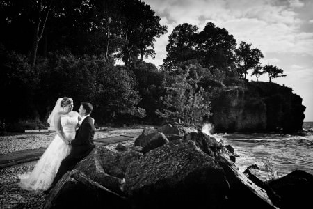 A black and white image of a bride standing and leaning into the groom's arms as he sits backs and rest on a large boulder on Catawba Island with large trees behind them and the shoreline of Lake Erie to their left.