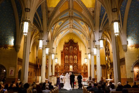 An image of a bride and groom standing at the altar with their best man and maid of honor in purple inside the Cathedral of St. John with cream colored pillars that form into beautiful arches on the ceiling which is painted blue with stars and the rest of the walls are a bright yellow with wooden accents.