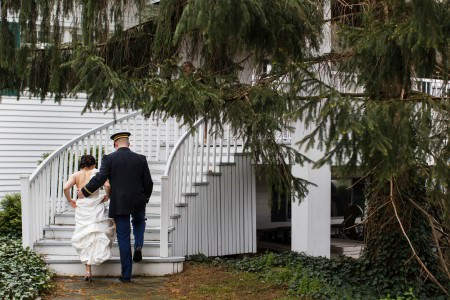 A image from behind of a bride in her white gown ascending the white rounded steps of the Chagrin Valley Athletic Club while her groom has his arm around her wearing his military dress blues assisting her with green pine trees in the foreground and ivy growing in the background.