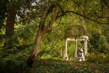 An image of a groom in a grey tuxedo is holding his bride in her white gown from behind while standing under a garden arbor with a wrought iron domed roof with white pillars surrounded by a forest of green foliage at the club at Hillbrook in Ohio.