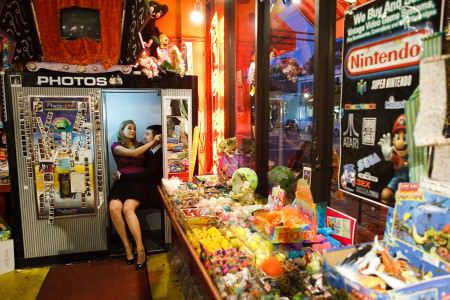 An image of an engaged couple where the woman is wearing a black skirt and purple top looking directly at the camera while sitting on the man's lap who is in a dark suit inside a photo booth which is part of an eclectic store full of bright colored trinket toys sitting on a table all in the right of the photo .
