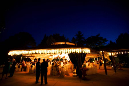 An evening photo taken at dusk of the outdoor tented space at Delucas Place where the reception tables and chairs are covered in white linen and the tent awnings are lit up with golden yellow lights and the guests are mingling in the chais and standing on the cement ground where the sky in the background is a stunning deep blue color.