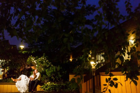 An outdoor evening picture of a bride leaning back into her groom's arms while they are sitting on a low wooden wall surrounded by green foliage on the walkway lit with soft yellow lights with a deep blue skyline and dark green leaves at a Doubletree hotel.