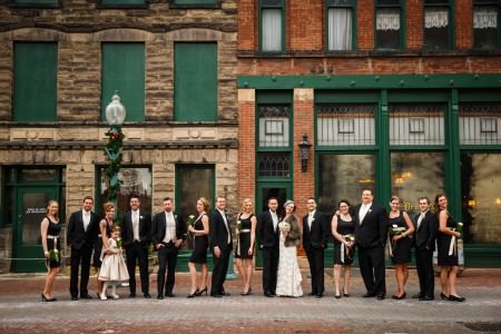 An image of a bride wearing her gown and a fur jacket posing with her groom in a black tuxedo along with their wedding party all in black with gold accents outside the landmark Bender's Tavern, a brick and stone building with dark green accents, on a December day in Canton, OH