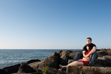 An image of an engaged couple where the woman is wearing a red dress and sitting snuggled in-between the man's jean-clad legs and black t-shirted arms on large rocks overlooking Lake Erie on the shores of Edgewater Park on a sunny and blue sky evening.