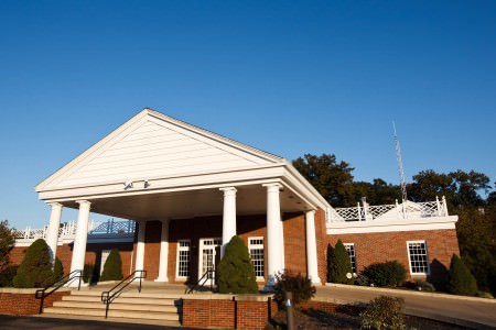 a picture taken outside of the red brick building of the Elyria Country Club where the entrance is in the middle left of the photo with it's white pillars and white overhang with cement steps in the lower left corner and dark green trees in the upper right corner all under a very bright blue sky.