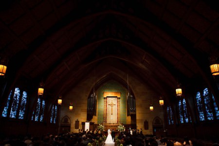 An image of a bride and groom standing facing the altar at the Fairmount Presbyterian Church where the chapel is darkened and the evening light is glowing blue through the stained glass windows and the altar is glowing with golden light where the ornate gold cross in the background is set up in front of a green wall.