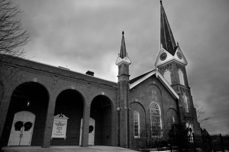 A black and white image of the outside of the brick Federated Church in Ohio with arched entry ways and two steeples on a dark and stormy day with bare trees and shrubs.