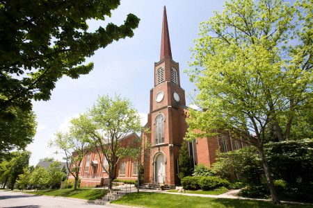 An image of a quaint red brick First Congregational Church in Hudson, Ohio with an arched white door under a tall steeple taken from the corner of the building where the church sits on the lower right half of the photo and the steeple extends up towards the top under a blue sky with tall green trees on either side of the photo.