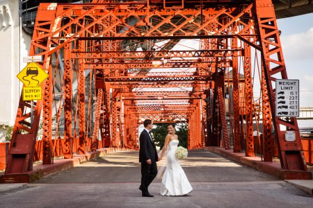 An image of a bride crossing the street of an orange industrial looking bridge holding her groom's hand and looking over her shoulder smiling at him while holding her white bouquet in the other hand.
