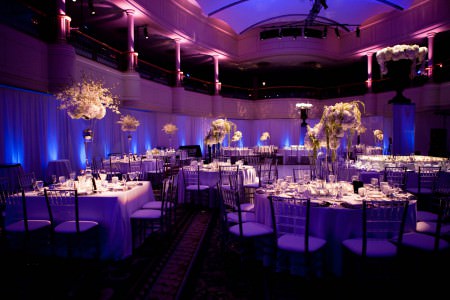 A wider view of a balcony circling a two story ballroom decorated for a wedding reception with both square and round tables, white linens, and tall ornate white and green floral arrangements, the walls draped in white material, and blue and purple uplights shining on the drapes and ceiling.