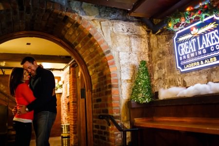 An engaged couple embrace in the left of the picture while standing under the arched brick entryway to the Great Lakes Brewing Company where the blue lit-up sign is mounted in the right of the picture to a stone wall.
