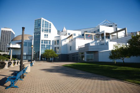 An outdoor shot taken of the back of the Great Lakes Science Center's white and glass multi-level building with a brick sidewalk and blue wrought-iron benches overlooking the water of Lake Erie on a sunny and blue sky day.