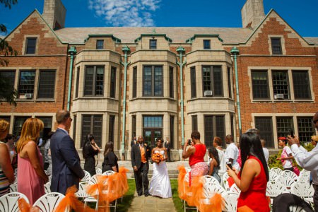 An image of a bride being escorted by her dad down the stone aisle with the brick and stone building of the Hathaway Brown School in the background holding her orange bouquet while the guests stand up watching from white folding chairs lined with large orange bows made from tulle.