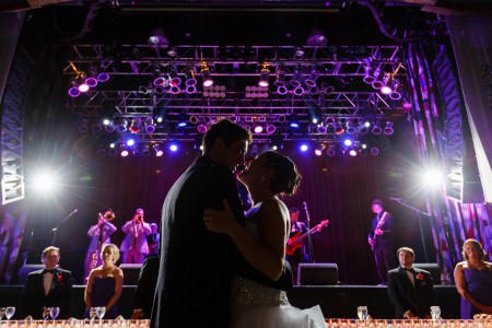 An image of a bride and groom slow dancing on the floor of the House of Blues in Cleveland with the head table in the background up against the stage where the band is playing under purple lights.