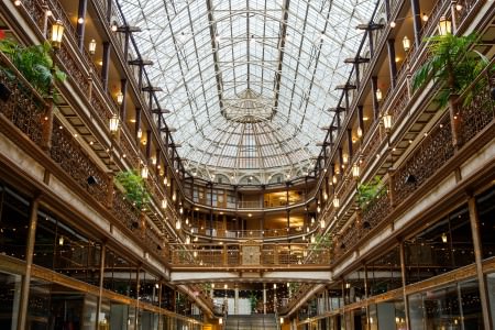 An image taken from the middle of Cleveland's oval shaped Arcade looking up from the ground floor towards the windowed ceiling and all of the levels of balconies with gold and wrought iron railings and cafe lighting draped around the room.