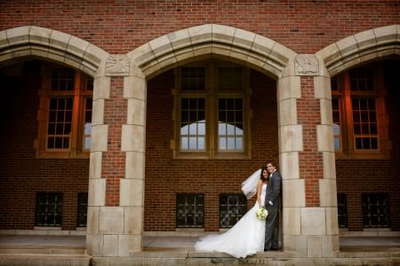 An image of a bride in her strapless white gown with her white veil blowing in the wind leaning into her groom in a dark grey tuxedo who is standing against a brick and stone pillar of the middle archway set in front of a building at the John Carroll University.