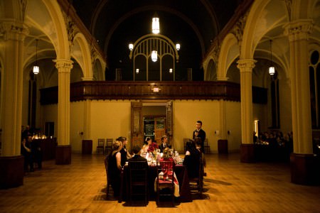 A wedding party is seated at a square head table set in the middle of the room at the Josaphat Art Hall with light colored ornate arched pillars and dark trim set around the room and a lighter colored wooden floor.