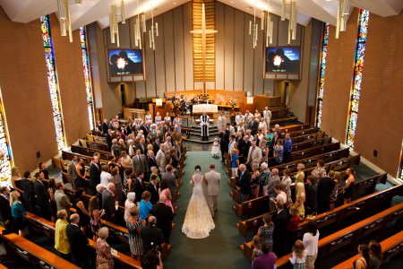 An image taken from the balcony of Kent United Methodist Church of a bride being walked down the green carpeted center aisle on the arm of her father heading towards the front of the sanctuary towards the groom and the waiting wedding party while the guests stand up from the wooden pews with brick walls and tall skinny stained glass windows on either side of the room.