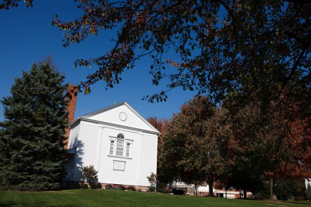 A picture of the small white Leroy United Methodist Church in the lower left of the photo sitting next to a tall pine tree on the left and taller maple trees on the right taken on a bright sunny fall day with under a brilliantly blue sky with leave from another tree hanging down in the upper right of the photo.