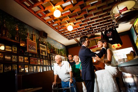 An image of a bride in her white gown sitting on a barstool looking up at her grown standing in front of her inside Luigi's restaurant in Akron, Ohio with wooden paneling and wooden ceiling beams.