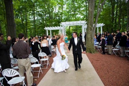 A picture of a bride and groom walking back down the outdoor aisle at Manakiki Golf Course in Willoughby, Ohio with guests standing from their seats on either wide while the couple is holding hands and laughing with a white arbor in the background under a canopy of tall green trees.
