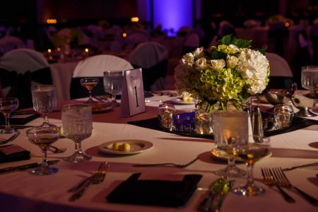 A close-up image of a round table set for a wedding reception labeled Table 1 with white linens and a green and white floral arrangement set in the center of the table while the rest of the tables in the background are out of focus set against a blue lit wall.