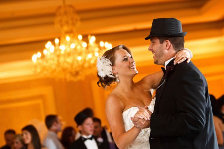 A waist high angled image of a bride in her white strapless gown with a feather in her hair smiling and dancing with the groom in his black tux with a formal hat inside a reception room glowing with a golden color at the McKinley Grand Hotel with a blurry chandelier in the background.