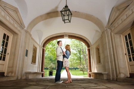 An image of a happy engaged couple facing each other and holding hands where the woman has on a short floral skirt and a jean shirt and the man has on jeans with a striped cream sweater standing in the center of the photo on a stone floor under the arched opening of a building at Miami University with green trees and grass beyond them through the opening.