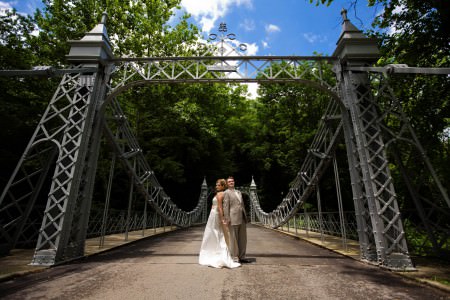 An image of a bride and groom standing in the middle of a bridge back to back but holding hands with their faces turned cheek to cheek looking at the camera surrounded by the wrought iron pillars and rails of the bridge with green trees in the background under a blue sky.