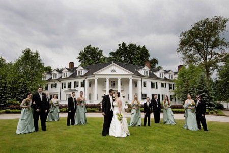 A bride and a groom stand on the green grass along with their wedding party spaced out a bit in front of the white pillared building of the Mooreland Mansion with green trees surrounding the building and grey cloudy skies above.