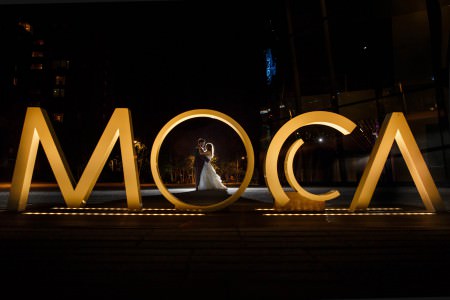 An image taken at night outside the Museum of Contemporary Art of a bride and groom embracing though the letter O of the large gold letters MOCA in downtown Cleveland, Ohio.