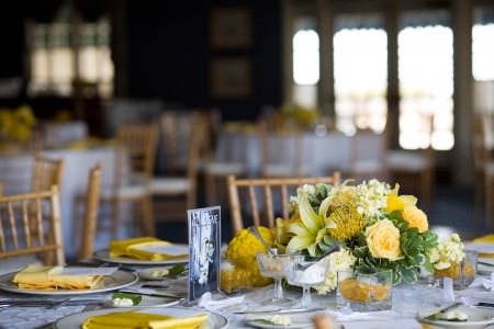 An picture of a room at the Oakwood Country Club decorated for a wedding reception with gold chairs and round tables set with white tablecloths and yellow linens as well as yellow and green floral arrangements where the table in the lower half of the photo is in focus and the rest of the room behind it is out of focus.