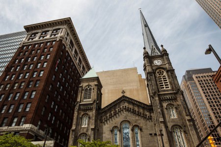 An outside picture of the front of the Old Stone Church in downtown Cleveland with the ornate stained glass windows and tall steeple nestled between other buildings under a blue and cloud streaked sky.