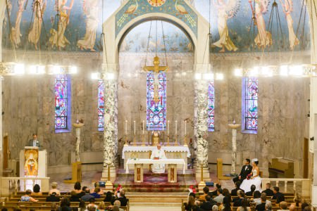 A picture taken from the balcony inside the sanctuary of Our Lady of the Peace church in Cleveland with marble pillars in a high vaulted ceiling with marble walls, beautiful purple and red stained glass windows behind the altar and a priest in robes standing at the table at the altar.