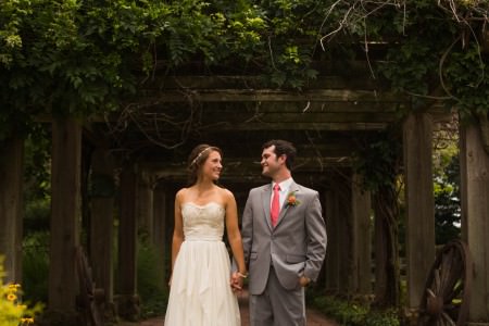 A bride in a strapless cream colored lace and tulle wedding gown is holding hands and turning her head to look at her groom in his grey tuxedo and bright pink tie while they stand under a wooden arbor with dark green foliage at Parker Ranch.