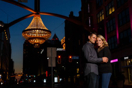 A picture of an engaged couple leaning into an embrace with their foreheads touching standing outside on the streets in Cleveland in jeans and dark shirts in the foreground of the left side while the stunning dark blue dusk sky sets in the background with the city lights on buildings and the Playhouse Square outdoor chandelier hangs with golden lights in the upper left corner of the background.