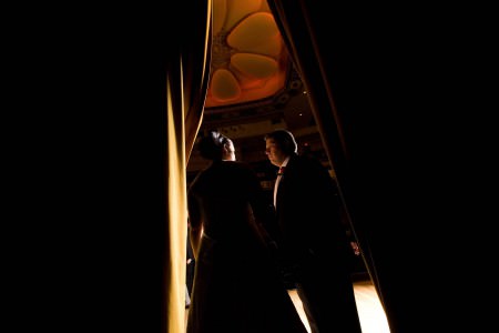 An image of a bride and groom standing face to face in the middle of the photo in the doorway of a darkened auditorium with their faces lit up and the image dark on either side of them at the Powers Auditorium.