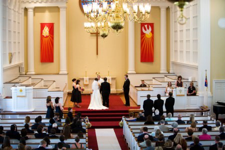 A bride and groom stand at the altar of a red carpeted church with yellow painted walls and white pillars along the back wall as well as white tiered pews on either side of the stage and red orange flags hanging from the walls as the wedding guests watch.