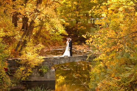A bride leans into her groom while turning her head towards the camera as they stand right in the middle of the picture which was taken outside on a fall day in Shaker Heights, Ohio standing on a stone path with a river running right through thick trees with leaves that have started changing from green to yellow and orange.