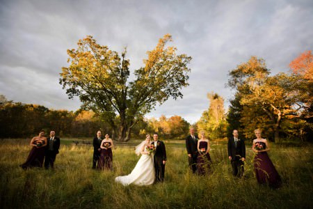 A fall outdoor image of a bride and groom standing in a grassy field under the historic Indian Signal Tree in Akron, Ohio with the bridesmaids wearing dark maroon gowns and the men wearing black tuxedos.