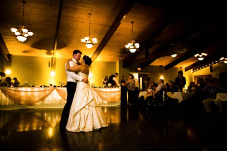 An image of a bride and groom slow dancing on the dark floor of the Skyland Pines reception hall with a wooden plank and beam ceiling lit up with golden yellow lights and the head table in the background with twinkle lights and tulle.