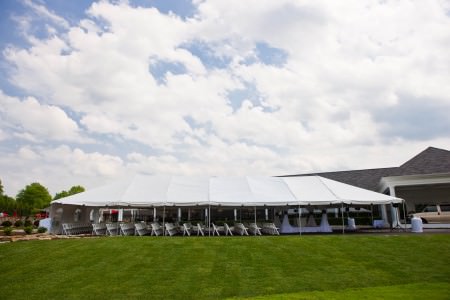 A picture taken of the outside of the Squaw Creek Country Club from the front green lawn with a large white tent in the very middle of the photo with white folding chairs lined up facing the right of the photo towards a square draped wedding altar all under a white puffy cloud sky.