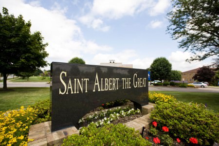 An image of a dark marble rectangular sign positioned in the middle of the photo with white lettering reading Saint Albert The Great set in the middle of a flower bed with green shrubs, and yellow, red, and white flowers in the middle of a green grassy lawn.