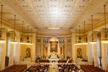 A photo taken from the balcony of St. Ann Church overlooking the large vaulted sanctuary that has cream colored walls and pillars with ornate paintings set in marble on the back wall of the altar and where the priest is standing in the center aisle while the wedding guests are seated in the pews and the bride and groom are seated to the right of the altar area.